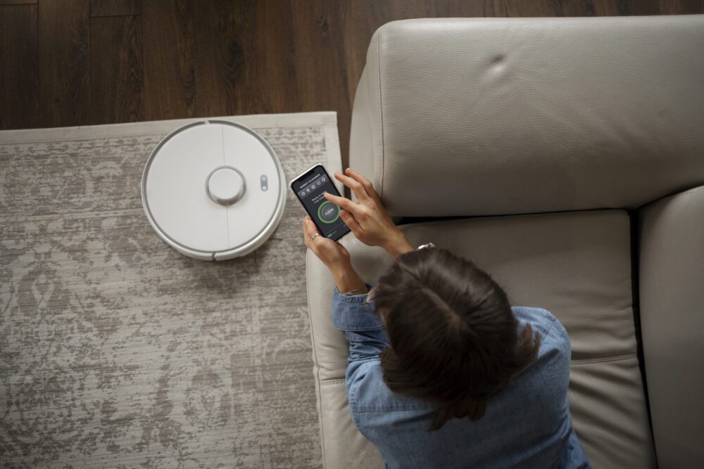 Kobieta leżąca na kanapie obsługuje za pomocą telefonu smart home uruchamiając odkurzacz